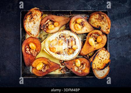 Im Ofen gebackener Camembert-Käse mit karamelisierten glasierten Birnen, Walnüssen und Trauben sowie Baguette auf rustikalem Metallteller Stockfoto