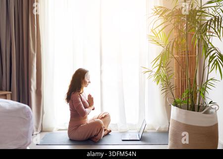 Erwachsene Frau übt sitzende Yoga-Meditation mit Laptop in der Nähe im Hotelzimmer Stockfoto
