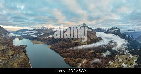 Blick aus der Vogelperspektive auf einen ruhigen See mit dem dramatischen Himmel, flankiert von herbstlichen Wäldern und schneebedeckten Gipfeln, Sils im Engadin/Segl, Graubünden, Schweiz Stockfoto