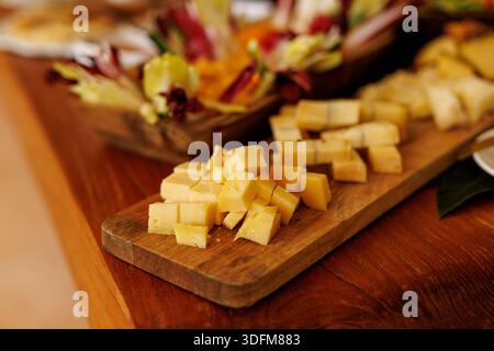Kleine Käsewürfel werden auf einem rustikalen Holzbrett mit Gemüseplatten im Hintergrund platziert. Stockfoto
