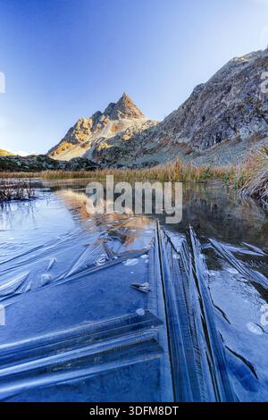 Blick auf einen teilweise gefrorenen See, der die zerklüfteten Gipfel unter einem klaren blauen Himmel reflektiert und einen atemberaubenden Kontrast aus eisigem Blau und warmen Bergtönen schafft. Stockfoto