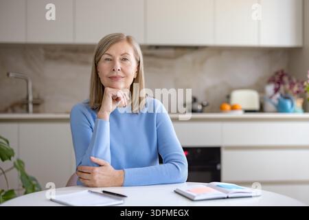 Eine ältere Frau in einem hellblauen Pullover sitzt mit einem Notizbuch an einem Küchentisch, blickt in die Kamera und plant den Ruhestand und ihre Zukunft mit ruhiger Zuversicht Stockfoto