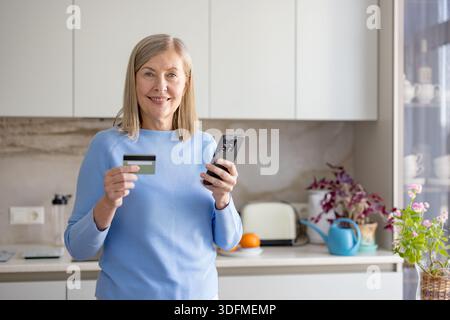 Seniorin in einer modernen Küche lächelt, während sie ein Smartphone und eine Debitkarte hält, und macht sichere Online-Banking- oder Online-Shopping-Transaktionen zu Hause problemlos und sicher Stockfoto