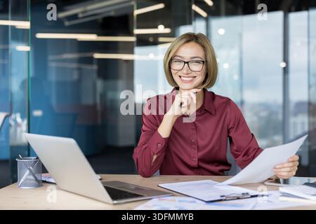 Eine junge Geschäftsfrau lächelt, trägt eine Brille und hält Dokumente in der Hand, während sie mit einem Laptop und Papierkram an ihrem Schreibtisch in einer modernen Büroumgebung sitzt Stockfoto