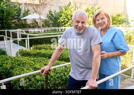 Seniorenmann mit Physiotherapeuten, der während der Rehabilitation im Freien geht und einen Gehrahmen zur Unterstützung und Erholung nach einer Verletzung oder Operation verwendet. Diese im Stockfoto