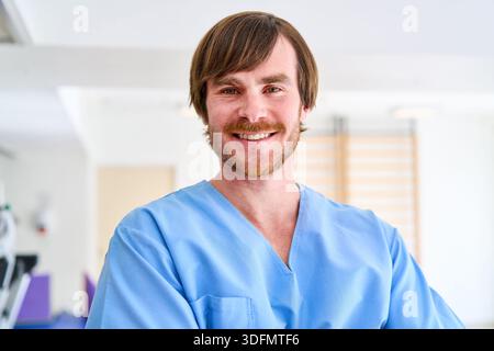 Porträt eines lächelnden männlichen Physiotherapeuten, der blaue Peelings in einem Rehabilitationszentrum trägt, zu dem der Arzt in einer Klinik bereit ist Stockfoto