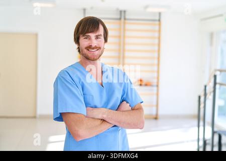 Ein Physiotherapeut in blauer Uniform steht mit überkreuzten Armen lächelnd in einem Reha-Zentrum. Dieses Bild kann für die Rehabilitation und Therapie im Gesundheitswesen verwendet werden Stockfoto