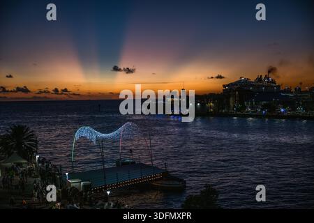 Der Blick auf die Uferpromenade leuchtet unter einem lebendigen Sonnenuntergangshimmel, wo Schiffe in der Nähe der beleuchteten Stadt ruhen und einen magischen Zauber über Willemstad, Curacao, ausstrahlen. Stockfoto