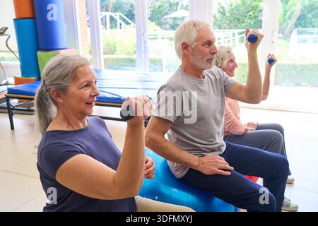 Ältere Erwachsene machen Kurzhantelübungen auf Übungsbälle in einem Reha-Zentrum. Sie arbeiten an Kraft- und Mobilitätstraining drinnen. Stockfoto