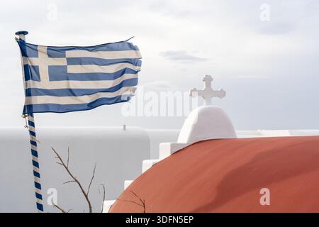 Blick auf eine griechische Flagge, die stolz gegen die weiß getünchten Gebäude mit roter Kuppel und Kreuz winkt, im Kontrast zum hellen Himmel, Mykonos, Griechenland. Stockfoto