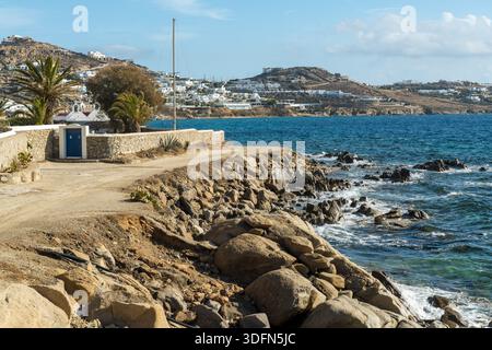 Blick auf die zerklüfteten Wellen, die gegen zerklüftete Felsen stoßen, ein sandiger Pfad, der an weißen Gebäuden auf einem entfernten Hügel, Mykonos, Griechenland, vorbeiführt. Stockfoto