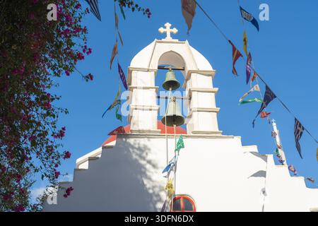 Blick auf die weiß getünchte Kirche mit einer markanten roten Kuppel, gekrönt von einem Kreuz, eingerahmt von lebhaften Bougainvillea und festlichen Flaggen, Mykonos, Altstadt, Griechenland Stockfoto