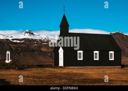 Der Blick auf die schroffe schwarze Kirche steht in scharfem Kontrast zum hellblauen Himmel und den schneebedeckten Bergen in der Ferne, Budir, Snaefellsness Halbinsel, IC Stockfoto