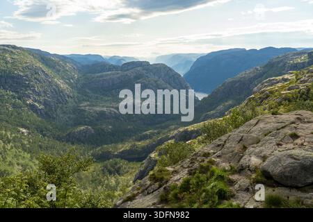 Blick auf smaragdgrüne Täler und zerklüftete Klippen, die in Richtung des ruhigen Fjords unter einem riesigen Himmel in Lysefjord, Rogaland, Norwegen, kaskadieren. Stockfoto
