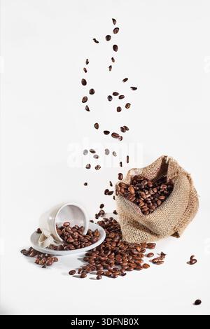 Geröstete Kaffeebohnen fliegen aus einem hessischen Sack, füllen eine Keramikschale auf einer Untertasse, natürliche Energie Stockfoto