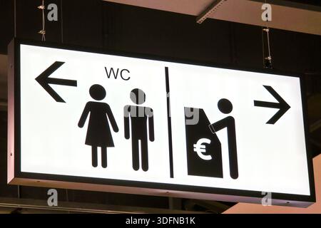 Beleuchtetes Schild für öffentliche Toiletten mit Piktogrammen für Männer und Frauen, Richtungspfeilen und einem Geldautomaten-Symbol in einem modernen öffentlichen Innenbereich Stockfoto