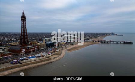Aus der Vogelperspektive auf eine Küstenstadt mit einem markanten Turm, Fahrgeschäften im Vergnügungspark, einem Strand und einem Pier, der sich unter einem bewölkten Himmel in Blackpoo ins Meer erstreckt Stockfoto