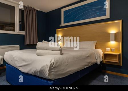 Ein Hotelzimmer verfügt über ein ordentlich gefertigtes Bett mit weißer Bettwäsche, ein hölzernes Kopfteil und gestapelte weiße Handtücher. Die Wände sind dunkelblau und der Boden ist Auto Stockfoto