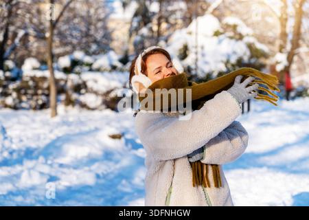 Junge Frau mit warmem Schal und Ohrenschützern, die draußen im Winterpark spazieren geht. Stockfoto
