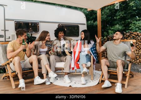 Interrassische Freunde, die Gitarre spielen und singen, amerikanische Flagge halten, Bier trinken, in den Sommerferien in der Nähe von RV sitzen Stockfoto