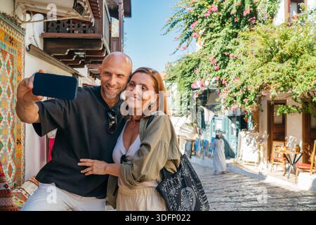 Ein Paar mittleren Alters lächelt beim Selfie mit dem Smartphone in der engen Straße im historischen Zentrum von Kas. Romantische Reise Liebe Beziehung, Honeymo Stockfoto
