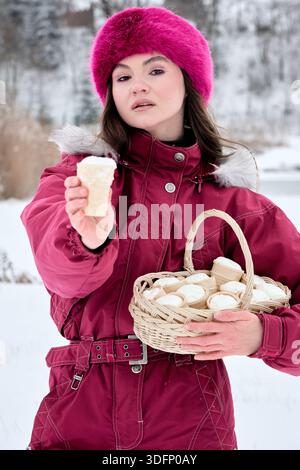 Dieses Stockbild zeigt eine junge Frau in einer Winterlandschaft, die Eiskegel aus einem bezaubernden Korb anbietet. Sie trägt einen rosa Pelzhut A Stockfoto
