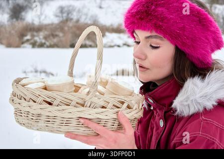 In einer fesselnden visuellen Erzählung steht eine Frau in lebendiger Winterkleidung inmitten einer ruhigen, schneebedeckten Landschaft, die einen gewebten Korb hält Stockfoto