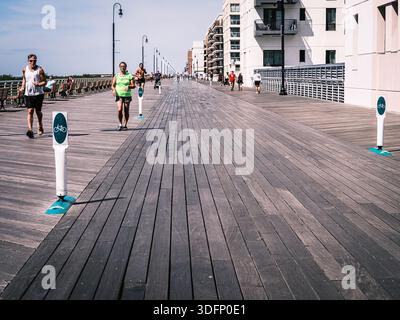 Die Menschen joggen und spazieren an einem sonnigen Tag entlang der Long Beach Promenade in New York mit Wohngebäuden, Bänken und offenen Flächen. Stockfoto