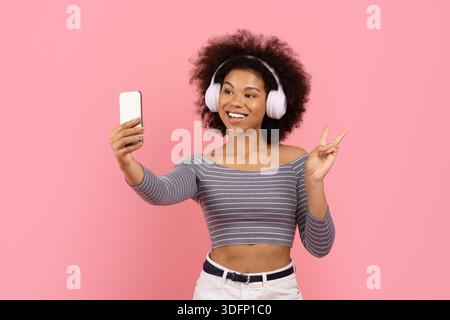 Fröhliche Frau, die Selfie mit dem Smartphone macht, während sie Musik hört, zeigt Friedenszeichen Stockfoto