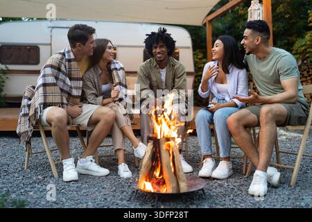 Multirassische Freunde sitzen in der Nähe von Lagerfeuer, reden miteinander, lachen, verbringen Herbstabend in der Nähe von Wohnmobil Stockfoto