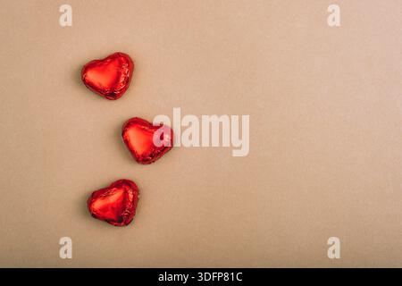 Drei rote Folie umwickelte Schokoladenherzen auf braunem Papierhintergrund mit Kopierraum. Stockfoto