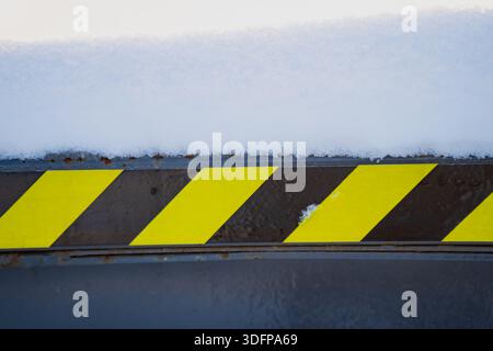 Schneebedeckte Stahlkante mit diagonalem Gefahrenstreifmuster in gelb und schwarz, Nahaufnahme der Kennzeichnung der Arbeitsschutzschilder auf der Ausrüstung im Winter l Stockfoto