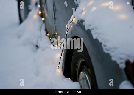Das geparkte Auto ist tief verschneit, mit leuchtender Lichtschnur um die Karosserie, Nahaufnahme des Hinterteils und des Rades in der verschneiten Einfahrt Stockfoto