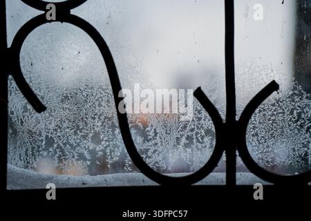 Frostmuster auf Fensterglas mit schwarzer schmiedeeiserner Silhouette, kalter Winterstruktur und unscharfem Hintergrund im Freien durch eisige Scheiben Stockfoto