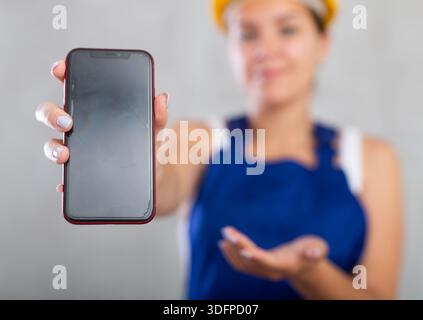 Nahaufnahme einer professionell lächelnden Ingenieurin, Bauleiterin in blauem Bauanzug und Sicherheitshelm mit Smartphone-Bildschirm Stockfoto