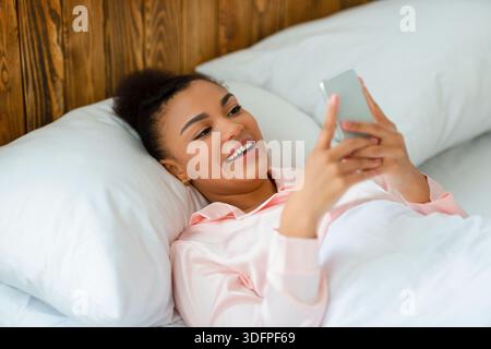 Mobiltelefonbenutzer. Positive Frau, die auf weißem Kissen im Bett liegt und SMS von jemandem liest und lächelt Stockfoto