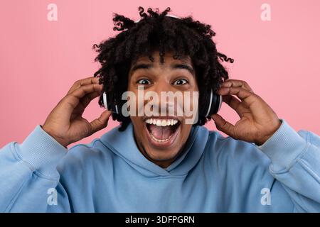 Ein überfröhlicher schwarzer Teenager genießt beliebte Musik im Headset und öffnet den Mund vor Spannung auf rosa Hintergrund Stockfoto