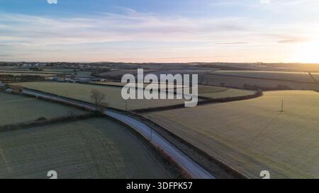 Aus der Vogelperspektive landwirtschaftlicher Felder und eine gewundene Landstraße bei einem frostigen Wintersonnenaufgang in Cornwall, England. Leichter Frost über Ackerland Stockfoto