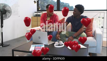 Zwei Männer, die im Wohnzimmer auf dem Sofa sitzen, lässig gekleidet, Laptop und rote Herzen sehen Stockfoto
