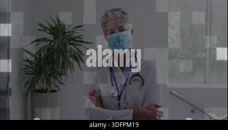 Stehender leitender Arzt, der die Arme im Klinikkorridor kreuzt, Laborkittel, Stethoskop, Maske, Ausweis trägt Stockfoto