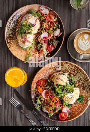 Hochwinkel-Flachbild eines dunklen hölzernen Cafétisches mit einer Vielzahl von Frühstücksgerichten, einschließlich Beerenpfannkuchen, frischem Gartensalat mit pochierten Eiern. Stockfoto