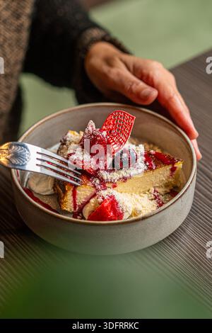 Nahaufnahme der Hände einer Person mit einer silbernen Gabel, um eine Scheibe cremigen baskischen Käsekuchen zu essen, der wunderschön mit frischen Erdbeeren garniert ist. Stockfoto