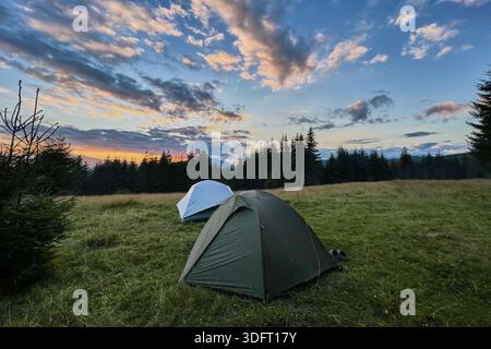 Wenige Touristenzelte in einem grasbewachsenen Feld am Abend, umgeben von Bäumen und Hügeln unter atemberaubendem Sonnenuntergangshimmel. Farbenfrohe Wolken verleihen der Szene eine schöne Note mit leuchtenden Orange- und Violetttönen am Horizont. Stockfoto