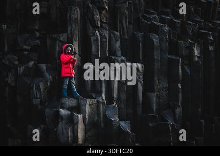 Der Blick auf eine einzelne Figur in einem leuchtend roten Fell steht auf den hoch aufragenden Basaltsäulen unter einem stimmungsvollen Himmel, ein markanter Kontrast zum dunklen Vulkangestein. Stockfoto