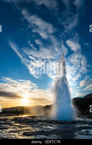 Der Blick auf den Geysir Strokkur erhebt sich in einer schillernden Darstellung vor dem goldenen Sonnenuntergang, mit Dampf steigt und trifft auf den lebendigen blauen Himmel, Haukadalur-Tal und Eis Stockfoto