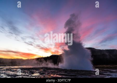 Der Blick auf den Geysir bricht in einem Ausbruch von kochendem Wasser vor dem Hintergrund feuriger Sonnenuntergänge aus, die den isländischen Himmel, Haukadalur Valley, Südwesten, malten Stockfoto