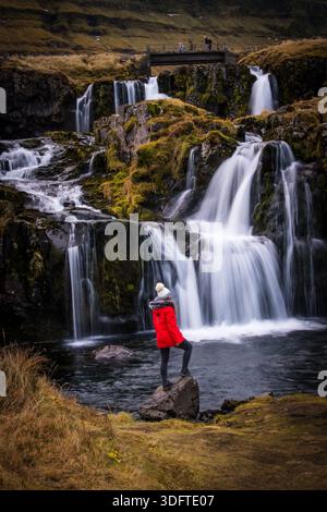 Blick auf kaskadierende Wasserfälle, die kraftvoll über dunkle Felsen fließen, während eine Person in einer roten Jacke kontemplativ auf einem Felsen, Grundarfjordur, West, steht Stockfoto