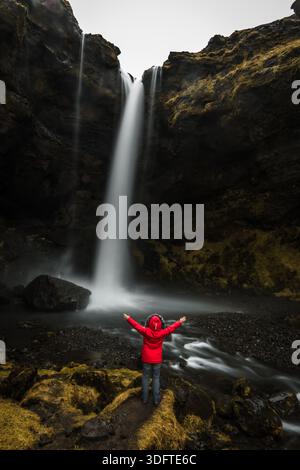 Blick auf eine einsame Figur in einer leuchtend roten Jacke, die vor dem ätherischen Kaskaden des Kvernufoss-Wasserfalls steht, ein krasser Kontrast zu den zerklüfteten Felsen Skog Stockfoto