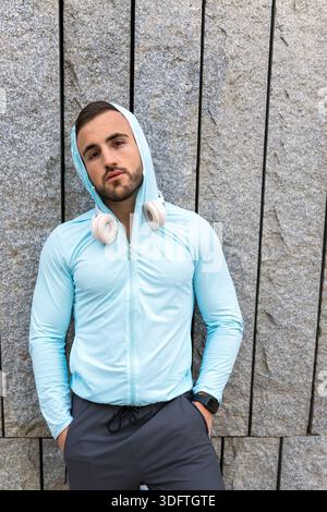 Der junge Mann steht selbstbewusst und trägt einen hellblauen Hoodie und Kopfhörer. Er posiert vor einer modernen Steinmauer und zeigt einen stilvollen urbanen Look und einen Stockfoto