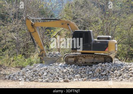 Gelbe Bagger-Baumaschine, die auf einem großen Steinhaufen aufliegt. Schwere Maschinen in Waldgebieten im Freien zeigen industrielle Leistung und Stockfoto